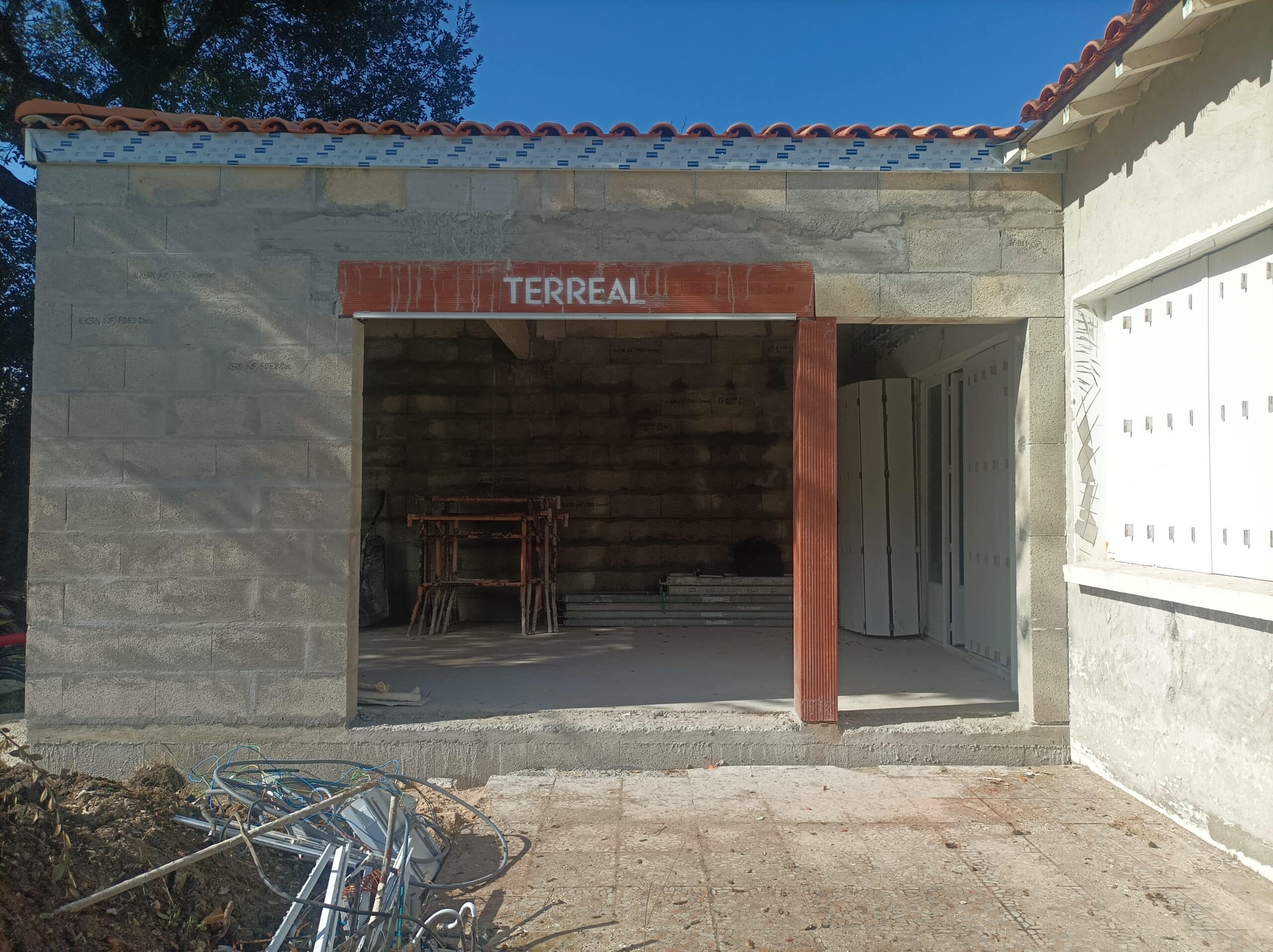 Construction d'un bâtiment en parpaings avec une ouverture de garage portant l'inscription 'TERREAL', sous un ciel bleu et des tuiles en terre cuite.