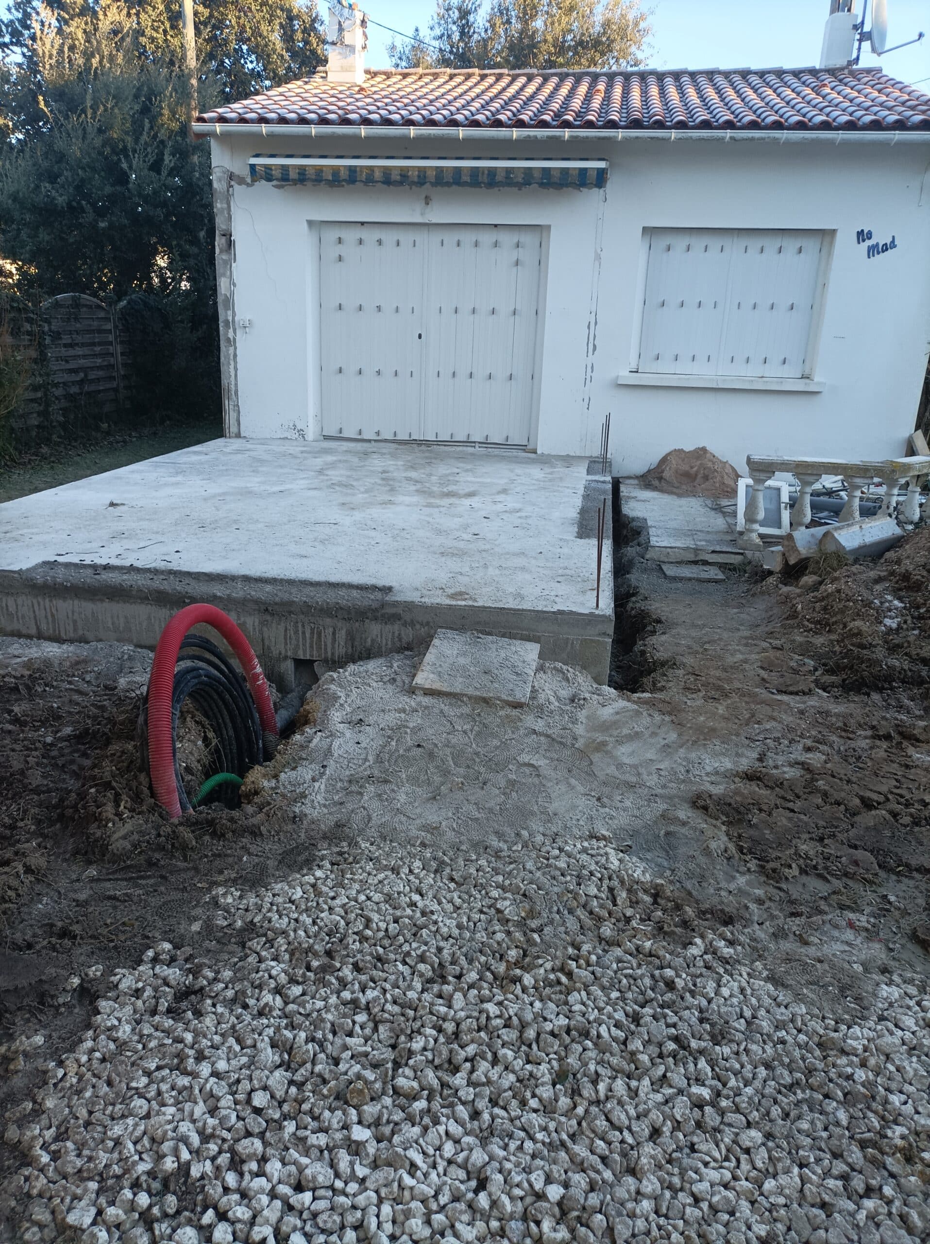 Chantier devant une maison blanche avec toit en tuiles, nouvelle dalle de béton, gravier et gaines électriques rouges et noires visibles.