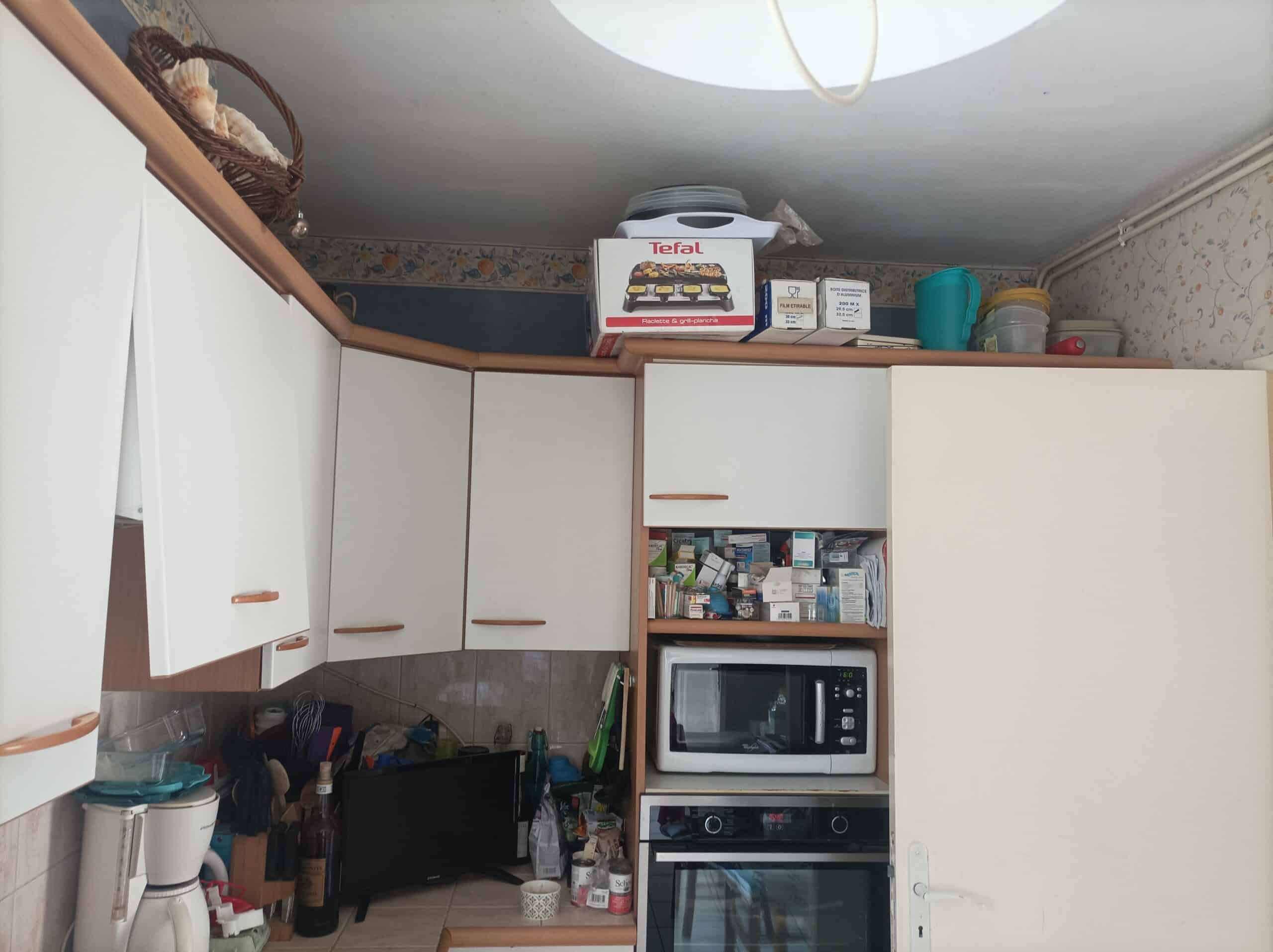 Cuisine avec placards blancs et bois, four encastré, micro-ondes Whirlpool, boîte Tefal, et médicaments sur une étagère ouverte.