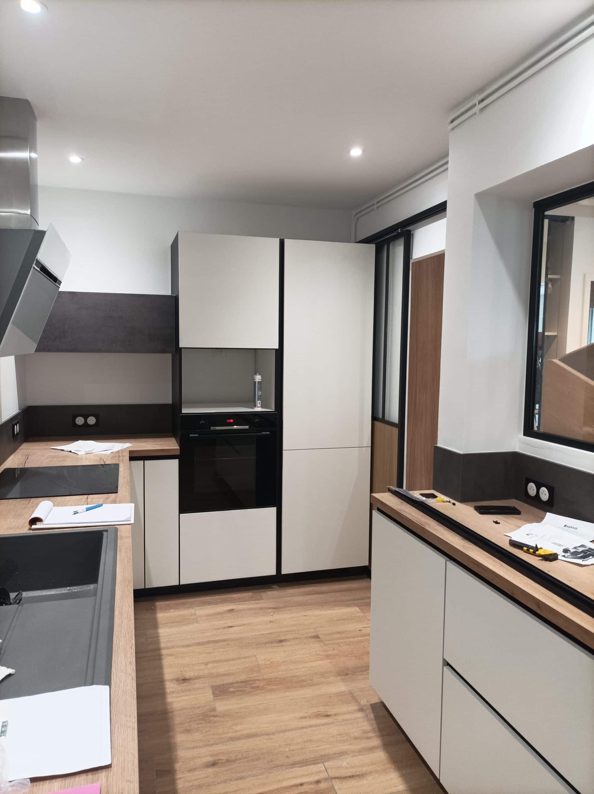 Cuisine moderne en cours d'installation avec meubles blancs, plan de travail imitation bois, électroménagers encastrés et sol carrelage imitation bois.