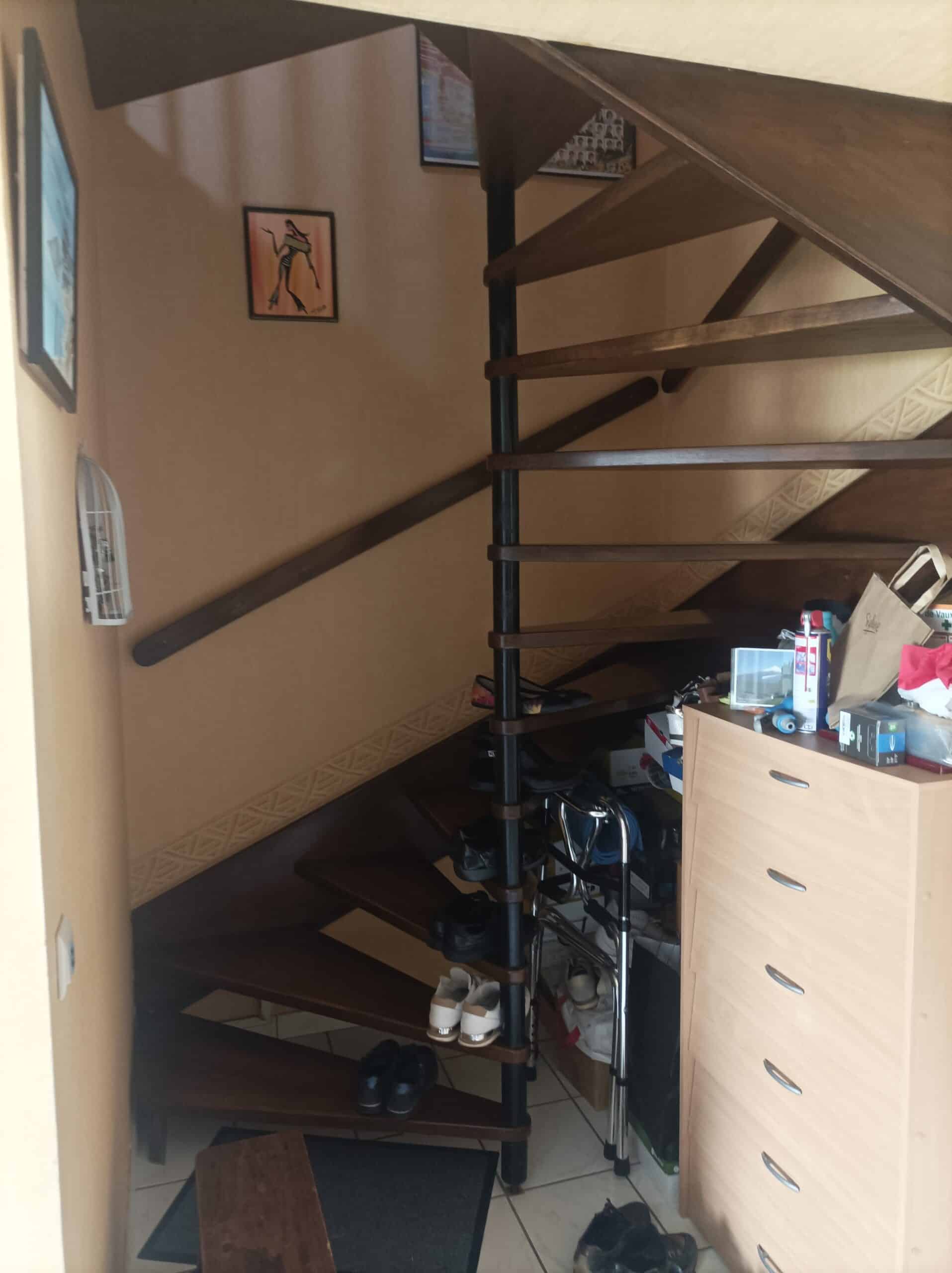 Entrée avec escalier colimaçon bois et métal, mur beige, chaussures sur les marches, et une commode claire encombrée.
