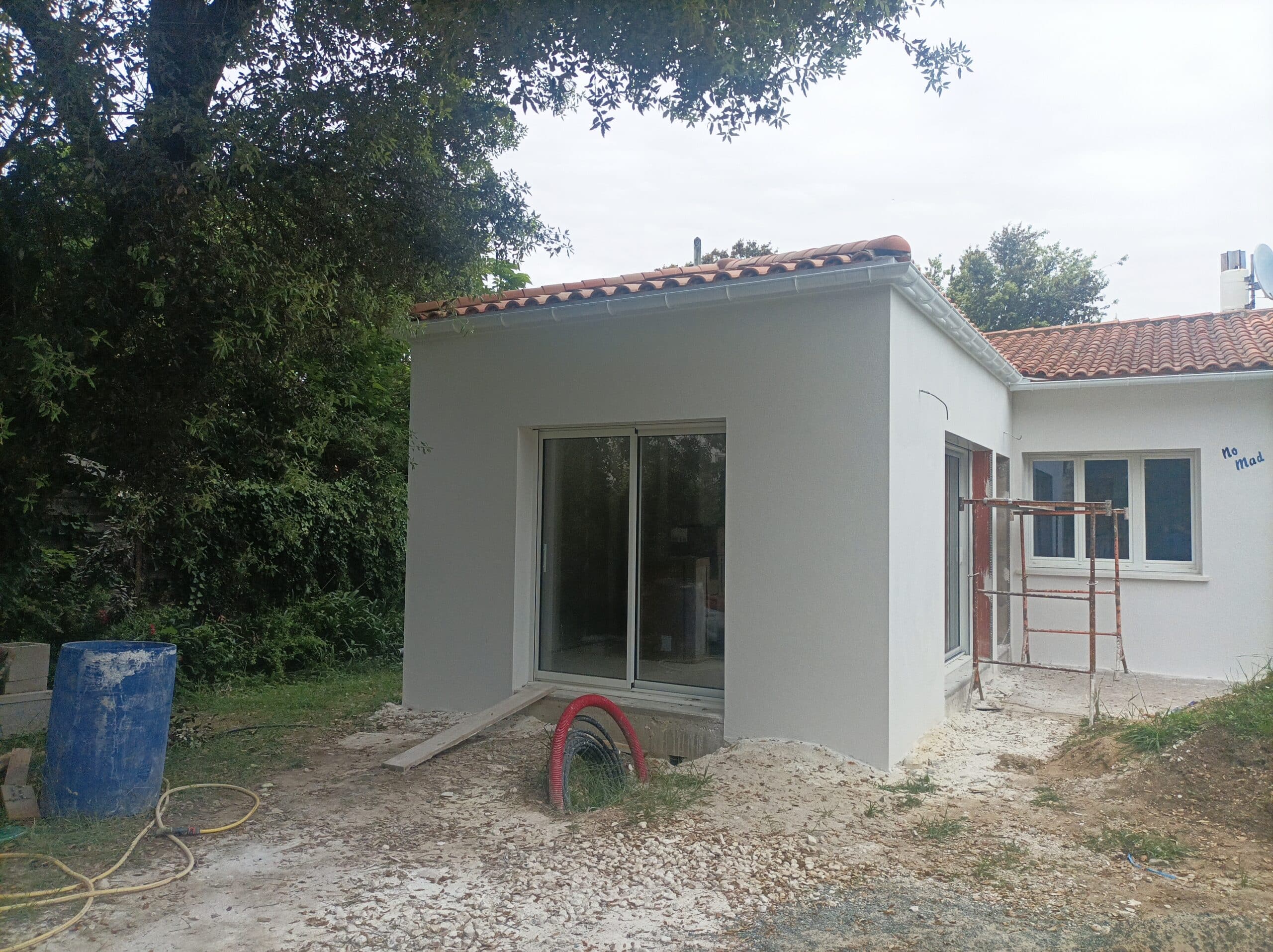 Construction d'une extension de maison aux murs blancs neufs, avec tuiles terre cuite, porte coulissante vitrée et échafaudage. Inscription "No Mad" visible.