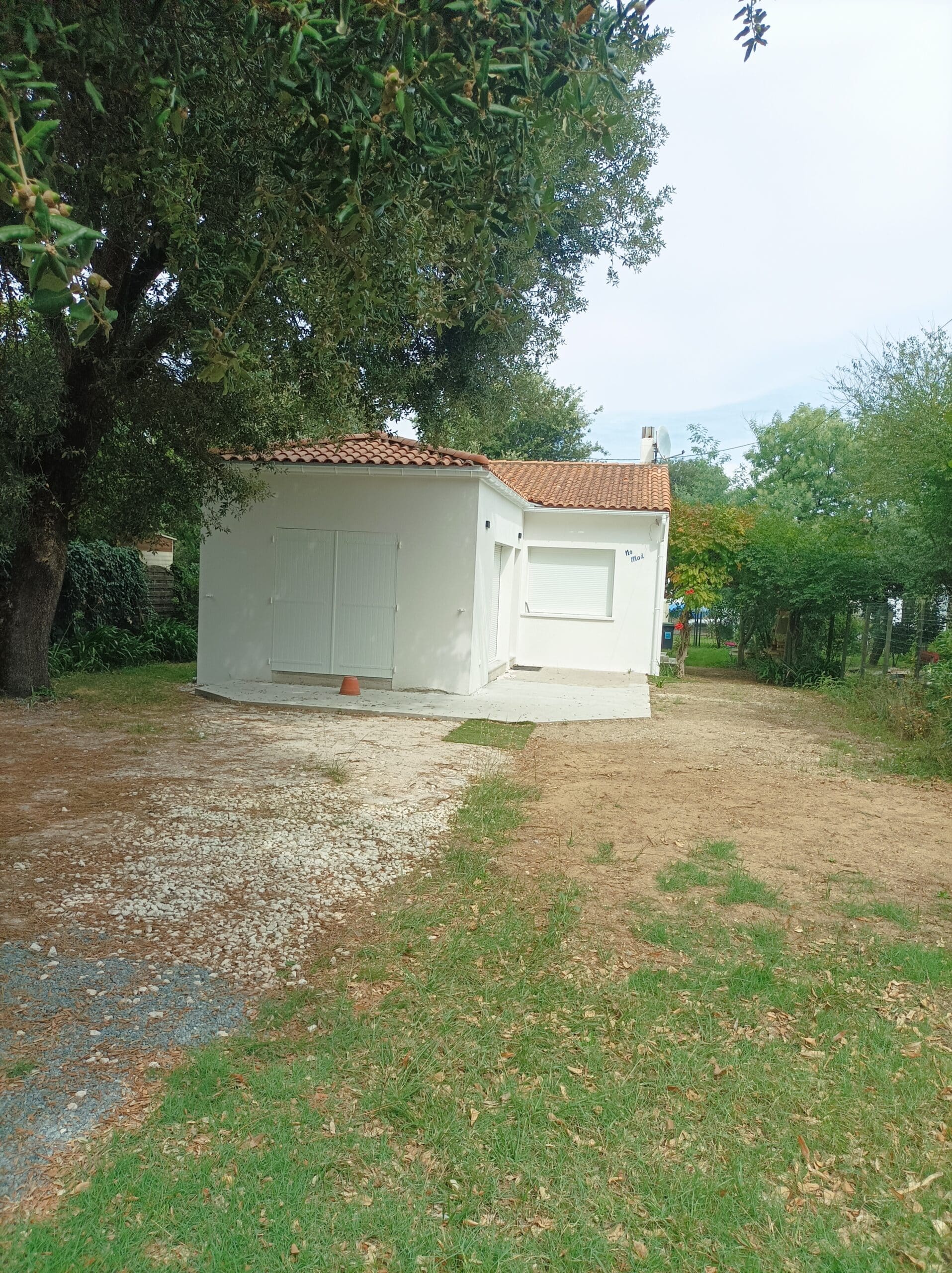 Petit bâtiment blanc neuf avec toit de tuiles rouges, entouré d'un grand chêne et de végétation, devant une allée de gravier.