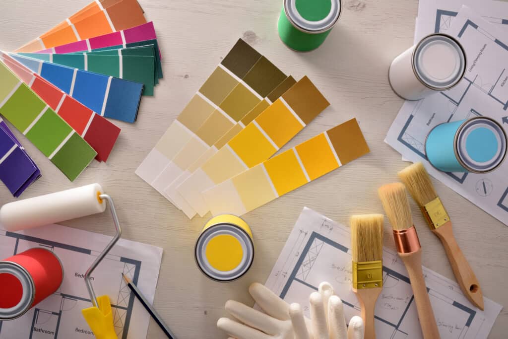 Choix de peinture et outils pour rénovation de maison Palette de couleurs, pots de peinture (rouge, jaune, bleu ciel, vert, blanc), rouleau, pinceaux et plans d'étage pour projet de rénovation intérieure.