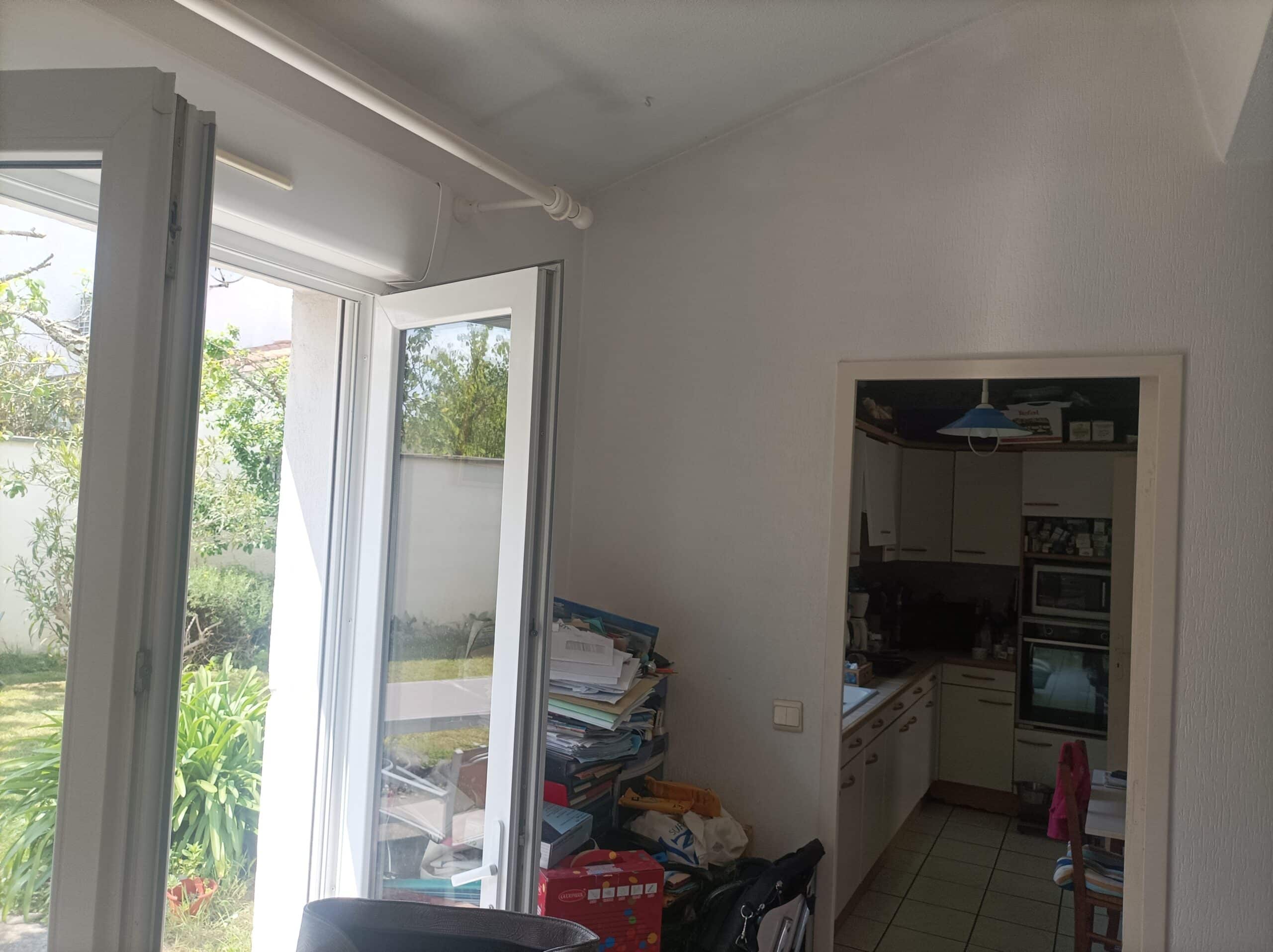 Intérieur lumineux avec porte-fenêtre ouverte sur un jardin verdoyant et une vue sur une cuisine en arrière-plan. Des piles de dossiers sont visibles.